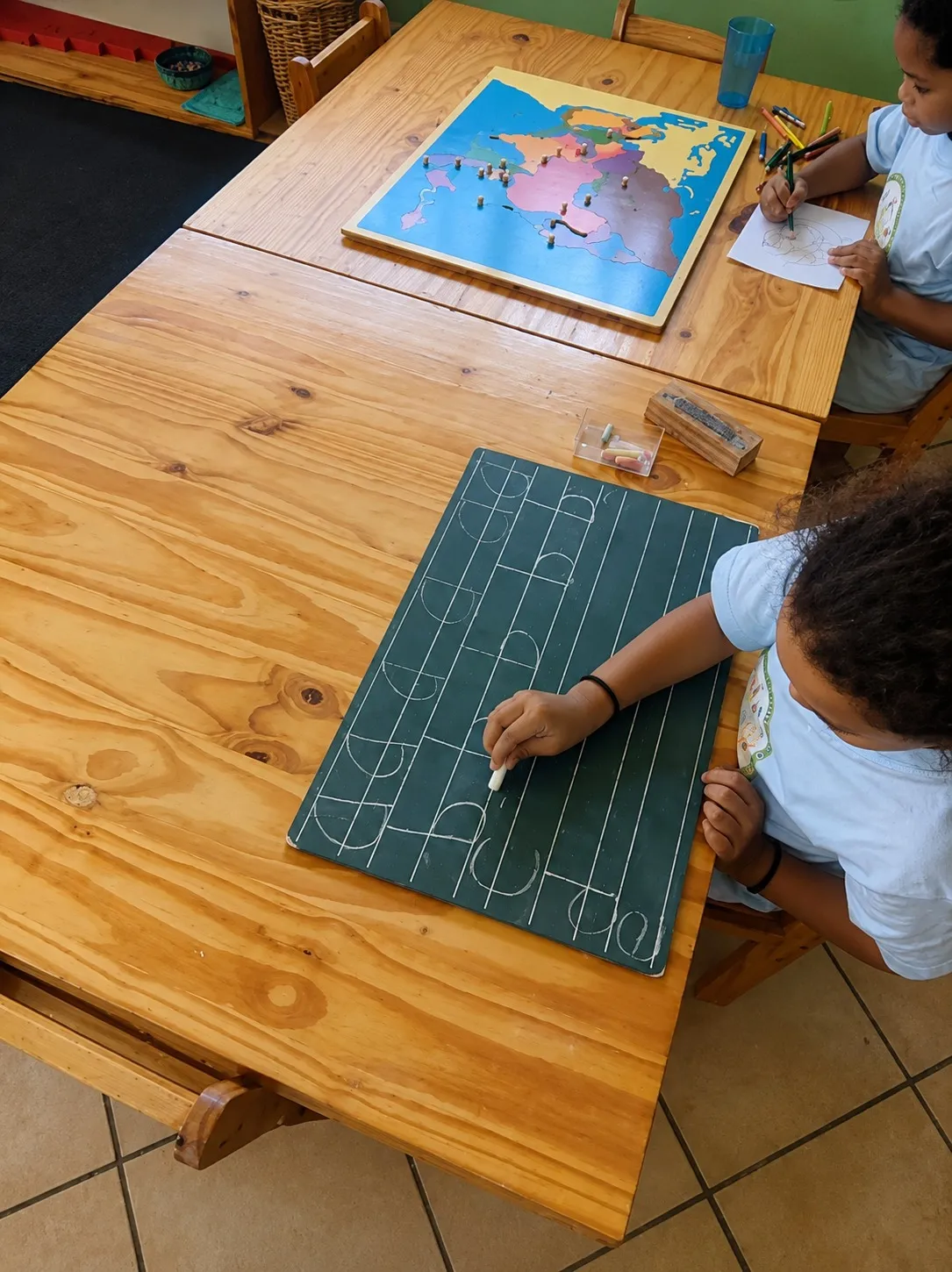 A child practising letter shapes on a chalkboard slate beside a world map puzzle